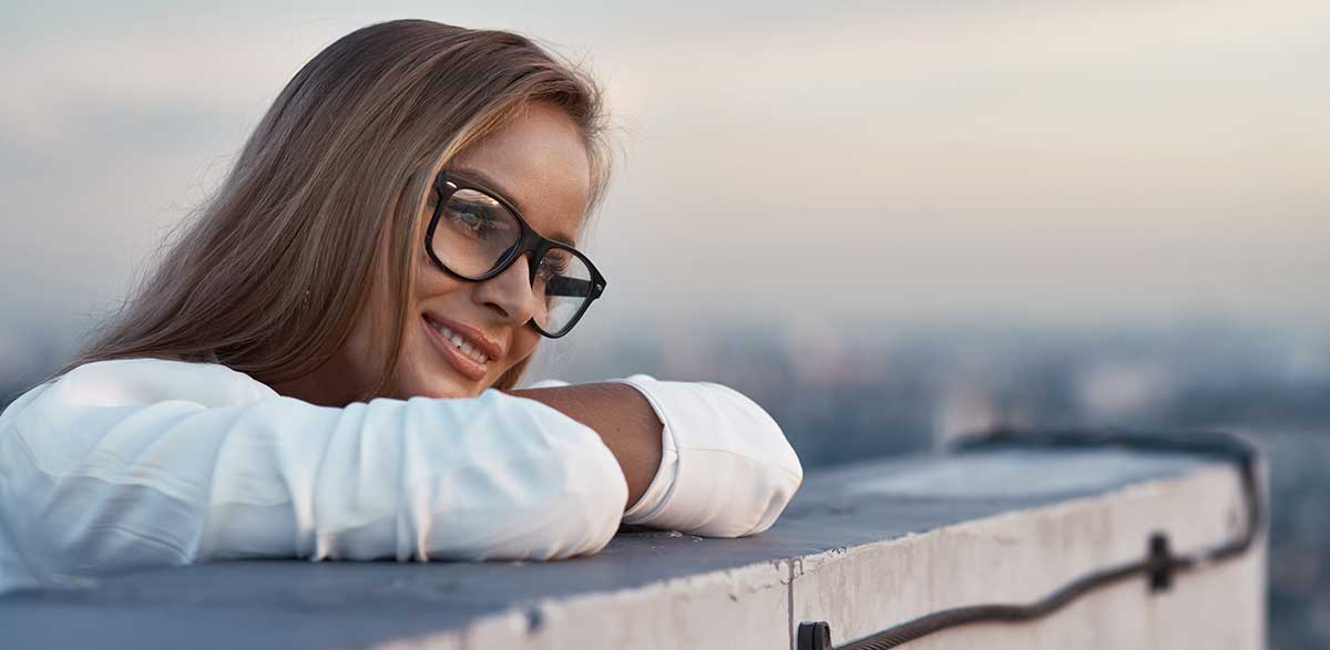 Portrait of a relaxed young businesswoman with glasses, standing on a balcony overlooking the city at dusk.